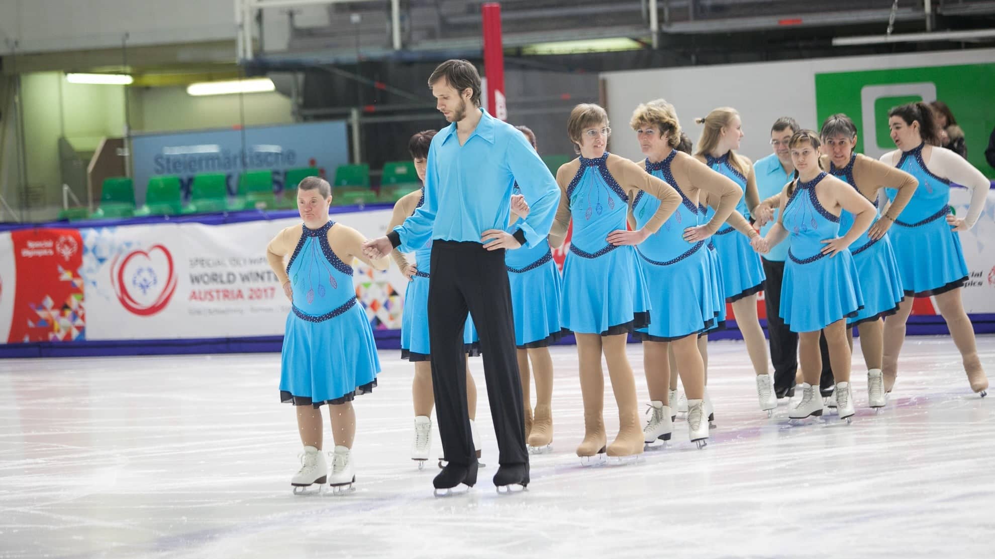 adaptive skaters all stand in a line as they perform on the ice. They are all wearing light blue dresses with a dark blue neckline and belt. One male skater wearing a matching long sleeve blue shirt and black pants holds the hand of the skater in the front of the line. 